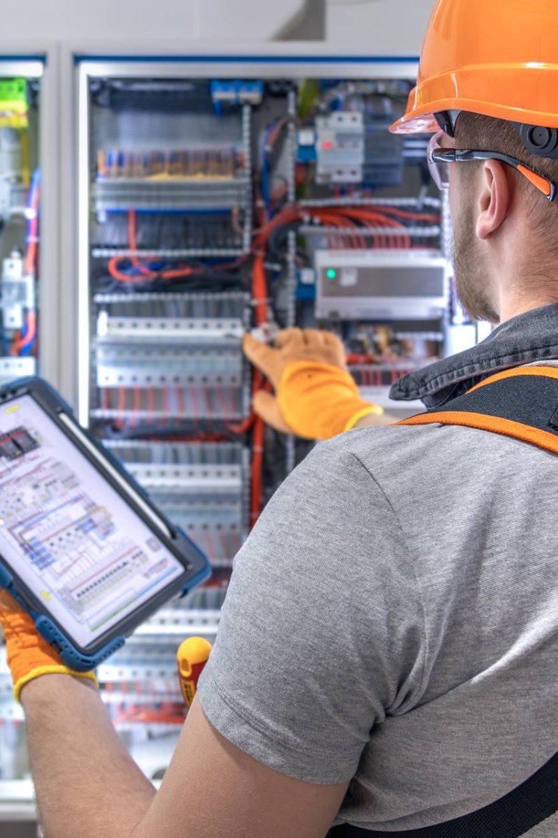 A male electrician in overalls, focused on work in switchboard with fuses, using tablet. High quality photo. An electrician with a tablet in his hand works in the switchboard. Copy space.